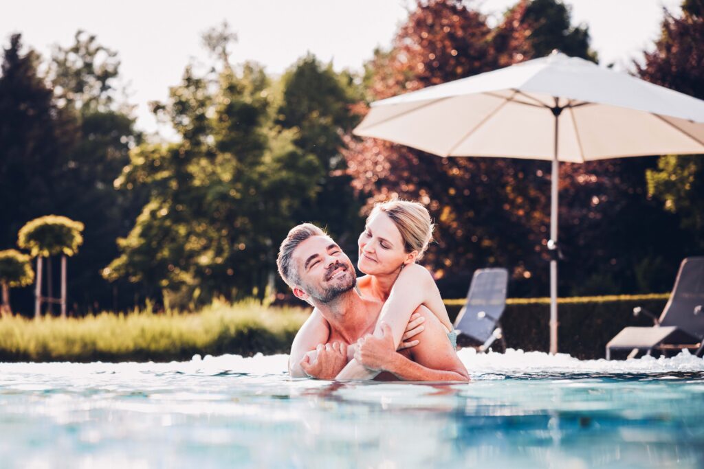 Ein Paar genießt die Zeit im Außenpool. Die Frau hängt über den Schultern vom Mann. Im Hintergrund Liegen und ein Sonnenschirm.