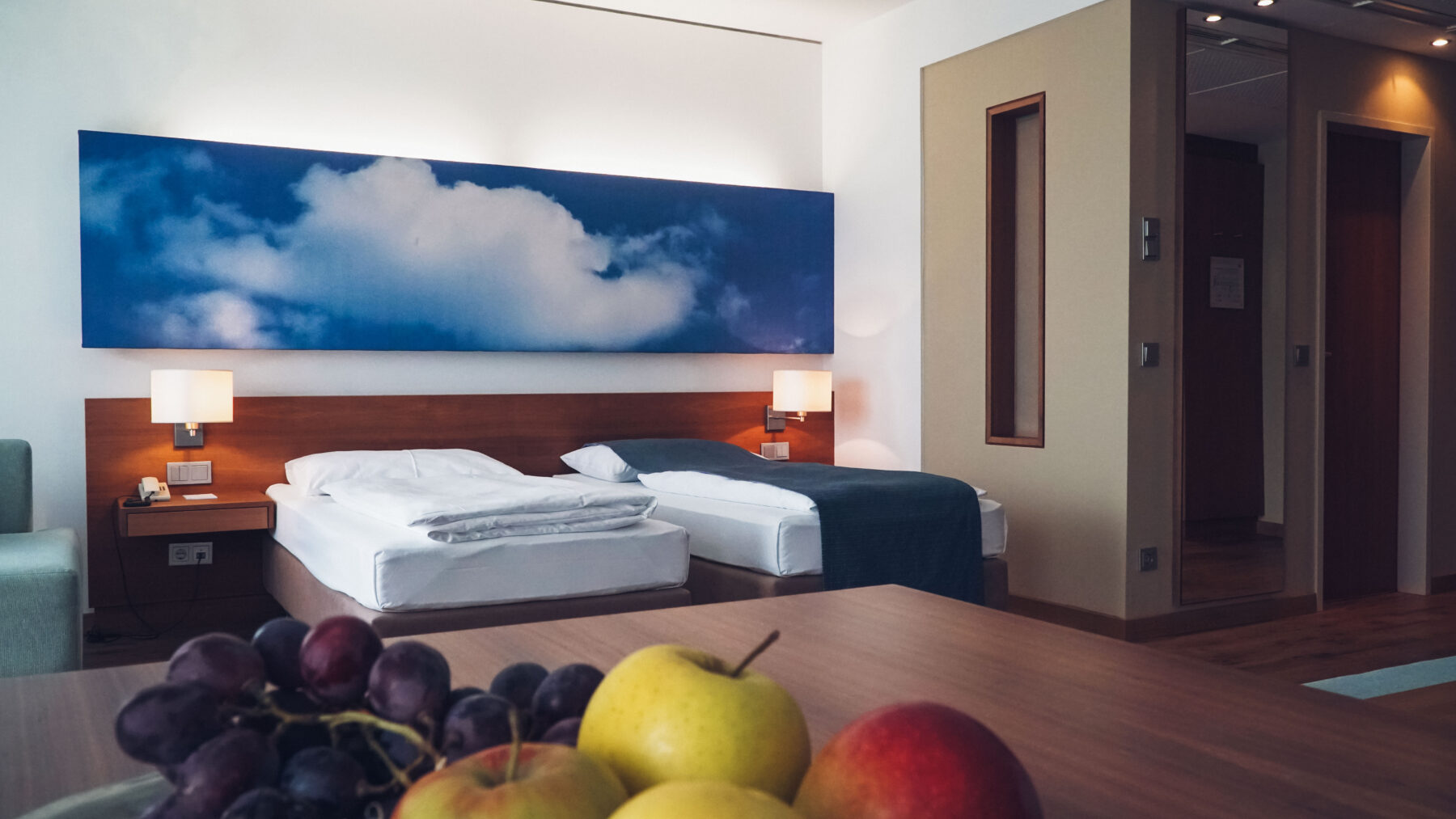 Hotelzimmer mit Doppelbett und einem Bild mit blauen Himmel und Wolken. im Vordergrund befindet sich eine Obstschale auf einem Tisch.
