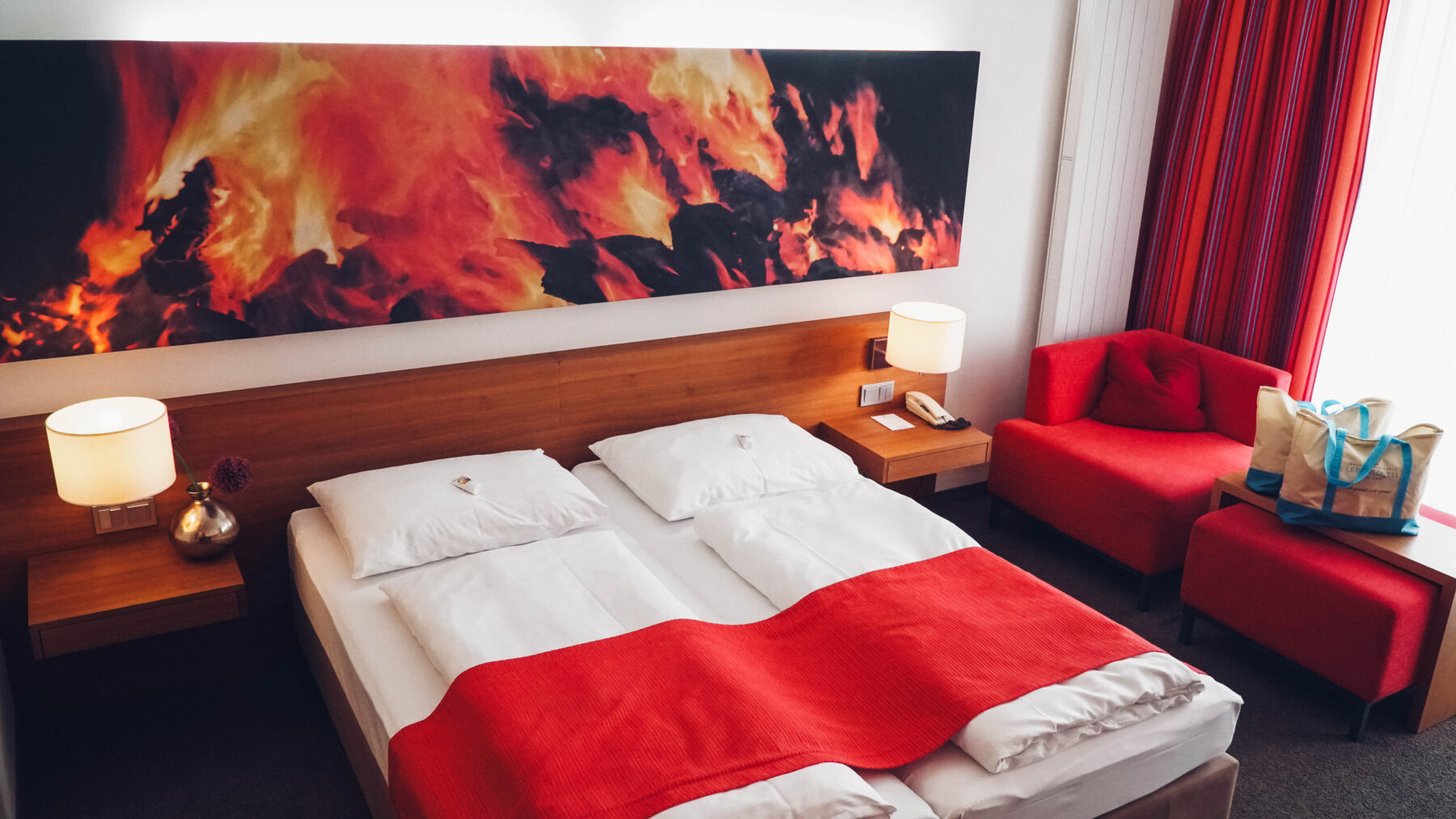 Hotelzimmer im Element Feuer - rote Akzente. Man sieht ein Doppelbett und einen Hocker und einen Couchtisch mit zwei Badetaschen. Auf dem Bild oberhalb des Bettes ist ein Feuer abgebildet.