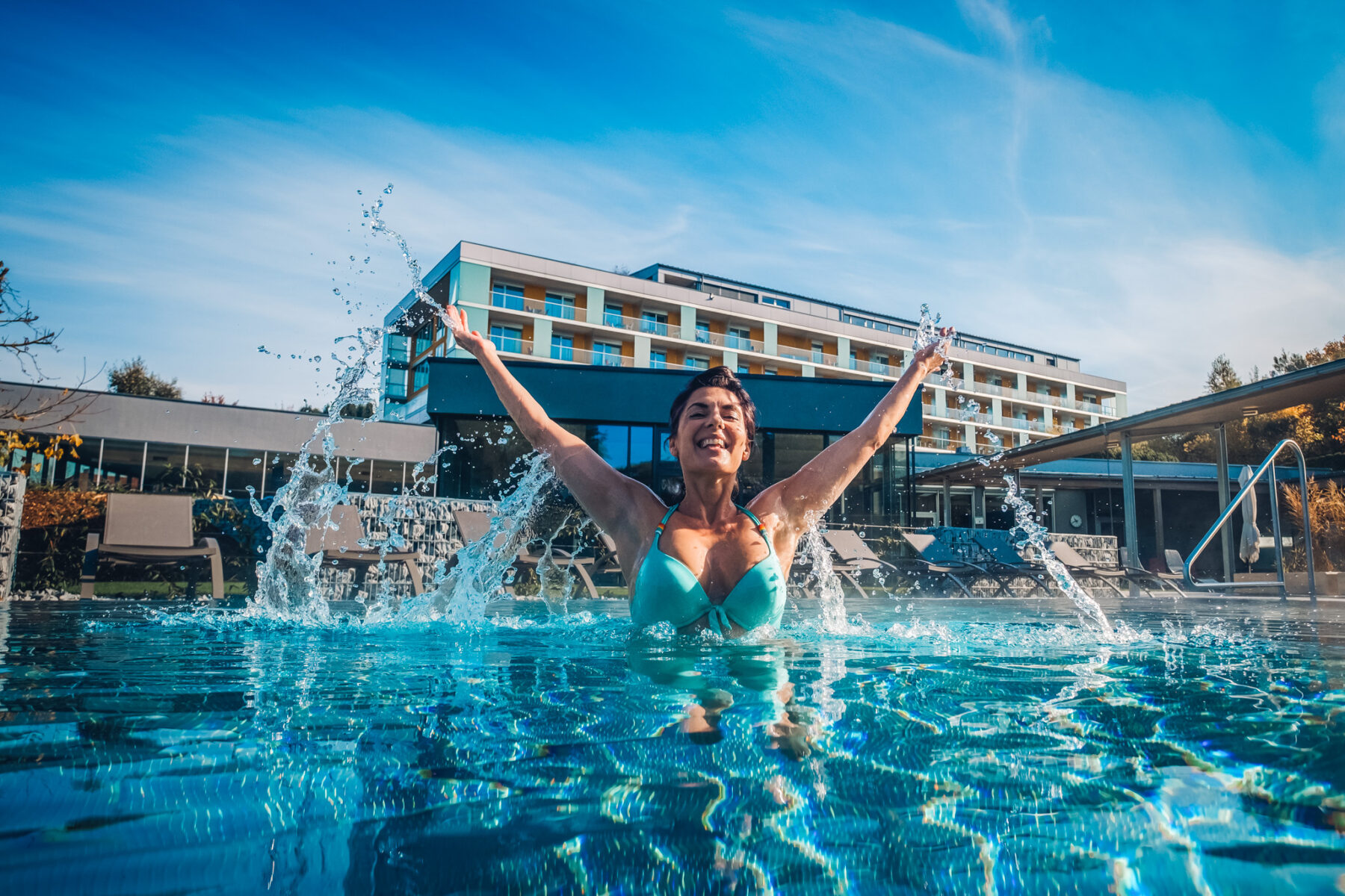 Glückliche Dame im Pool, welche vor Freude die Hände in die Luft hält und dabei mit dem Wasser spritzt. Im Hintergrund sieht man das ****s Hotel Lebensquell Bad Zell.