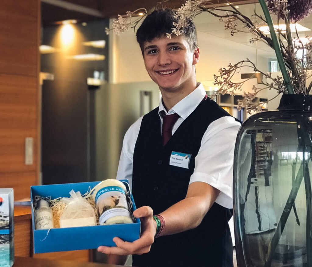 Man sieht einen Lehrling zum Hotel- und Gastgewerbeassistenten, welcher eine Geschenkbox in der Hand hält und diese präsentiert.