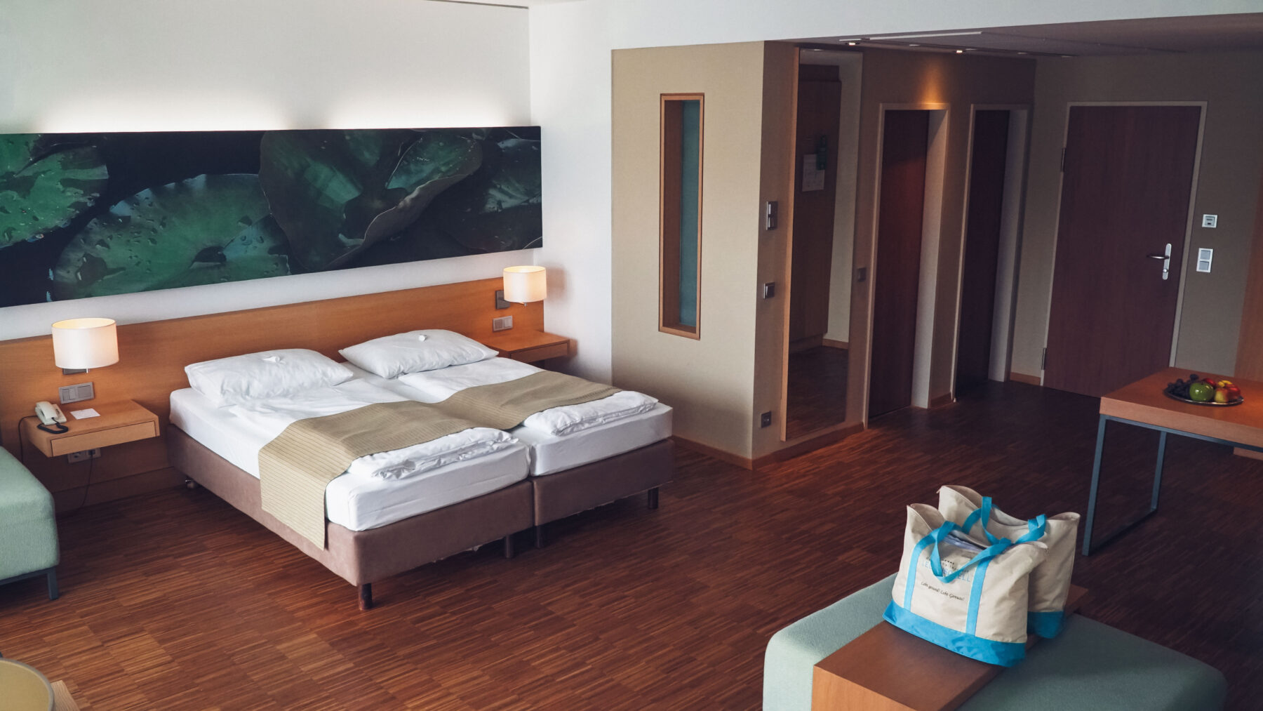 Hotelzimmer mit Doppelbett und Tisch. Am Couchtisch stehen zwei Badetaschen. Türen die zum Bad, WC und Zimmertür führen.