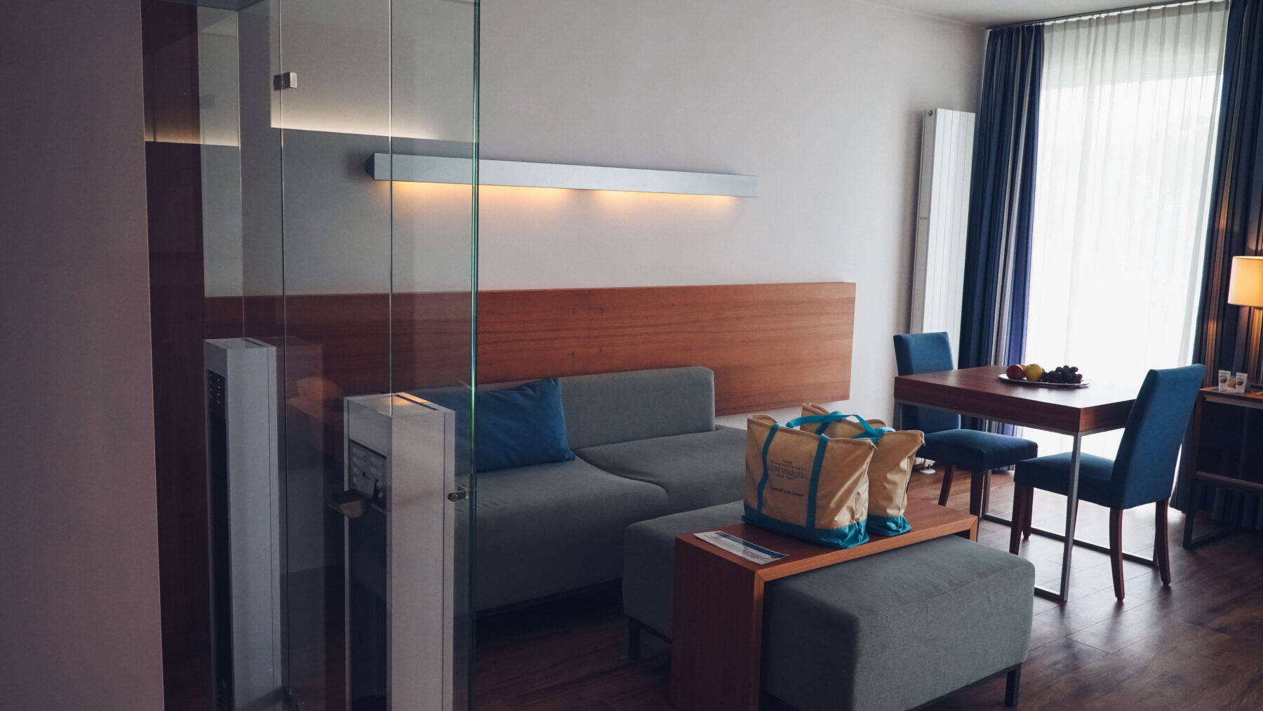 Hotelzimmer im Element Luft- blaue Akzente. Tisch mit zwei Sessel, kleine Couch und Couchtisch mit zwei Badetaschen.