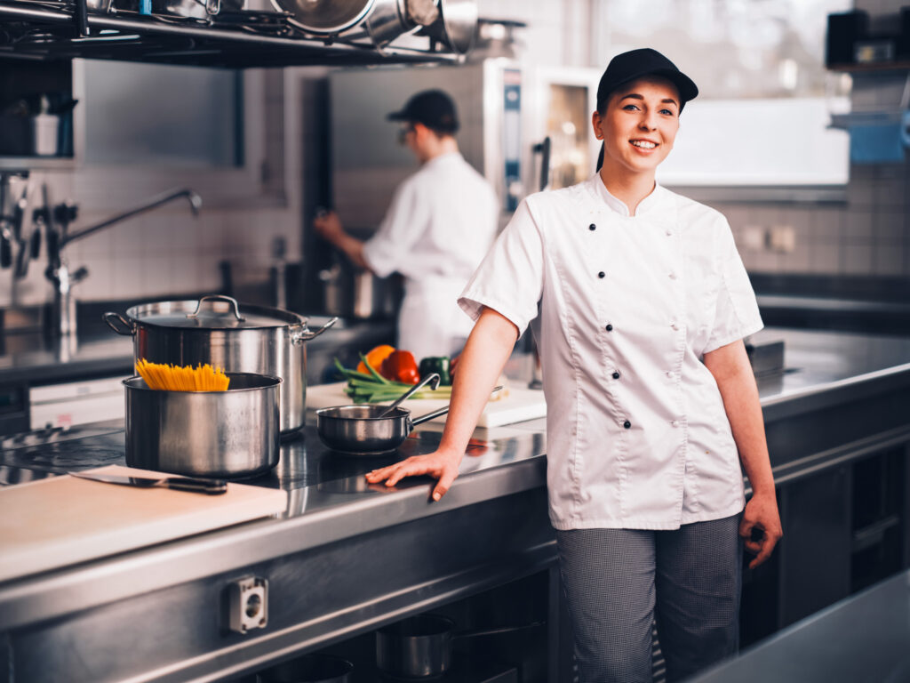 Im Vordergrund sieht man eine Köchin in der Küche, welche glücklich ist und neben sich Lebensmittel stehen hat. im Hintergrund ist ein zweiter Koch am arbeiten.