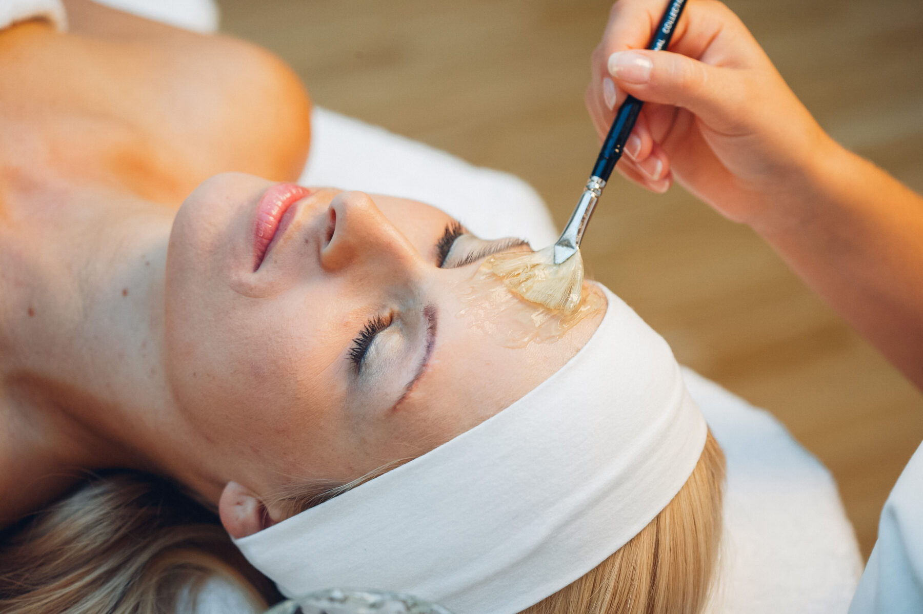 Kopf einer Dame. Auf der Stirn wird mit einem Pinsel eine Creme aufgetragen. Die Haare sind mit einem Band nach hinten gelegt.