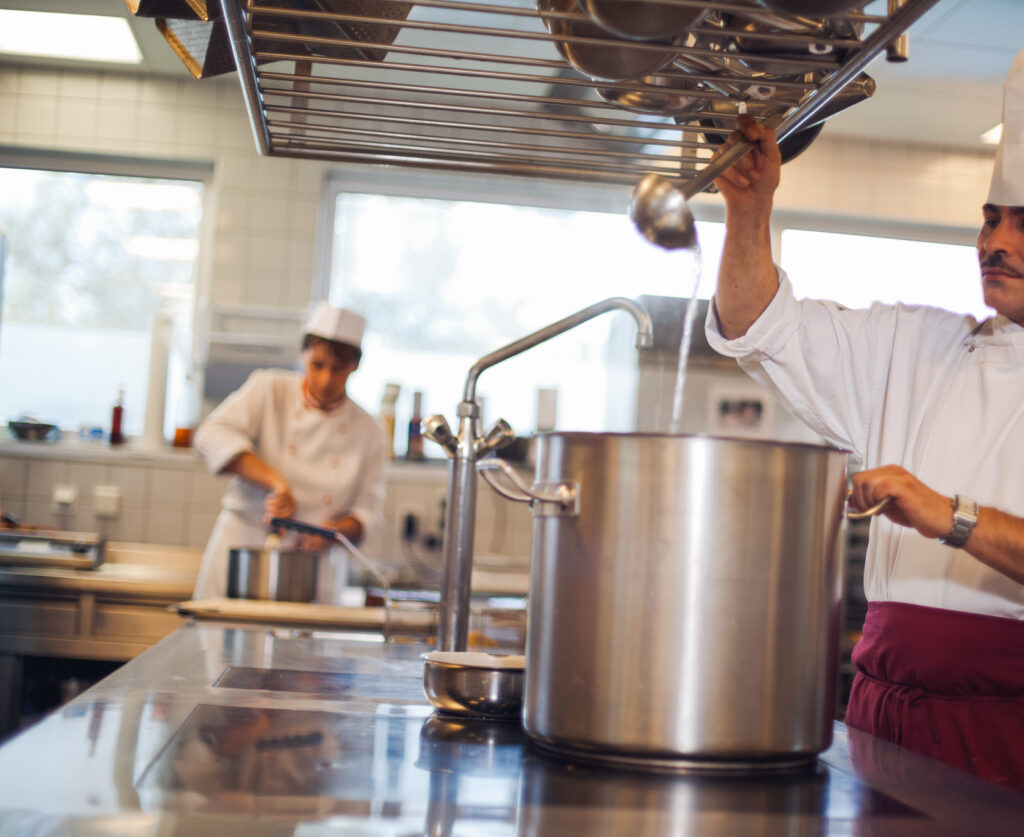 Man sieht eine Küche. Im Vordergrund ein Koch bei einem Kochtopf mit Kochlöffel in der Hand und im Hintergrund eine Konditorin am arbeiten.
