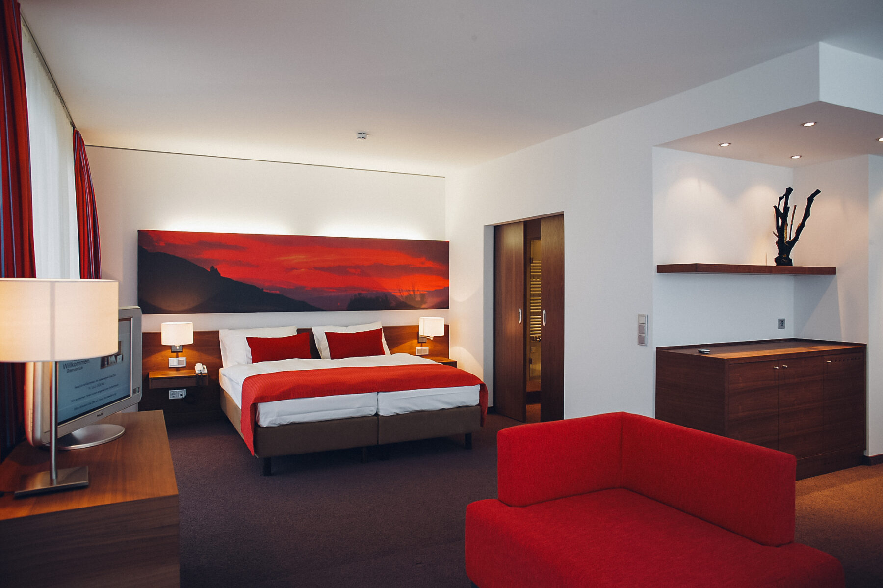 Lebensquellsuite mit roten Akzenten welches das Element Feuer weidergeben soll. Man sieht ein Bett, Couch, Fernseher und eine Tür Richtung Bad.