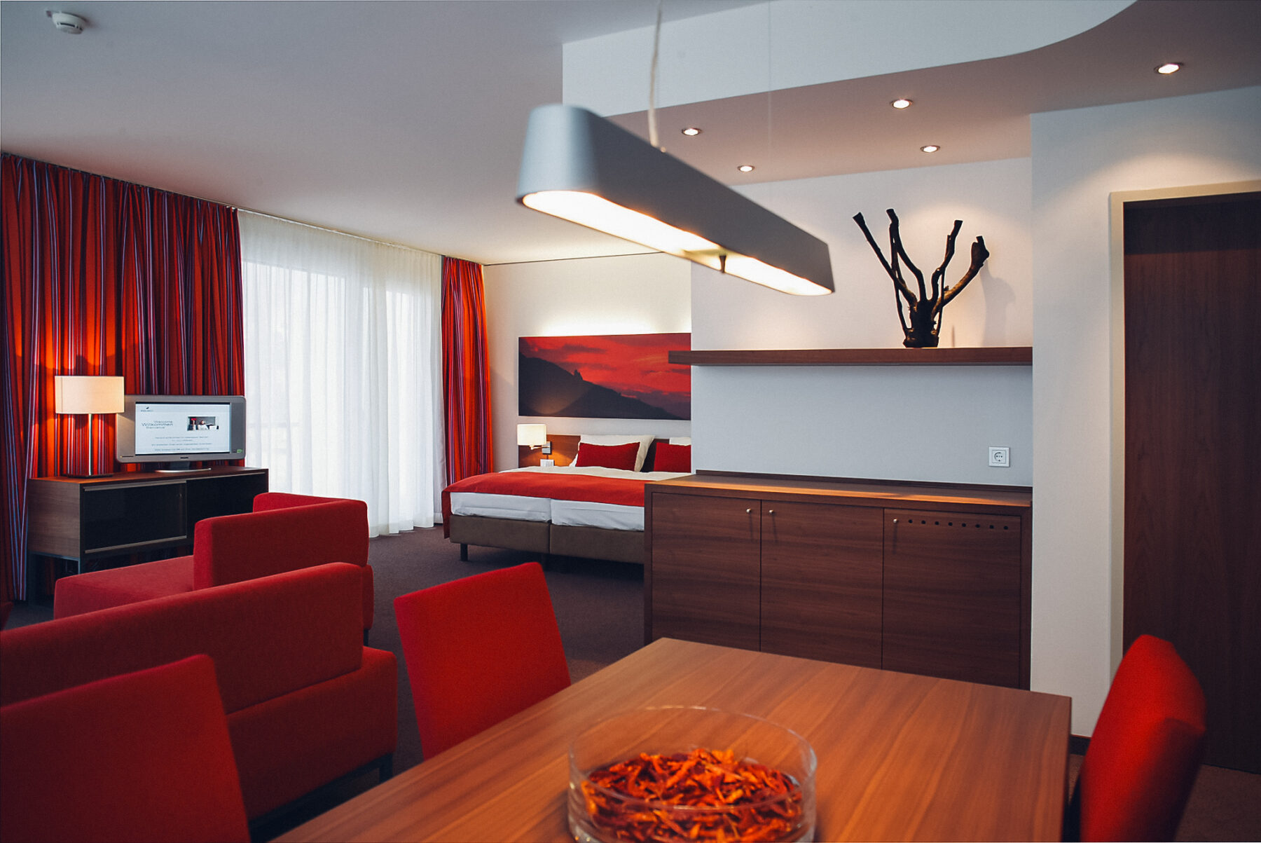 Lebensquellsuite mit roten Akzenten welches das Element Feuer weidergeben soll. Man sieht ein Bett, Couch, Fernseher, Tisch und Sessel