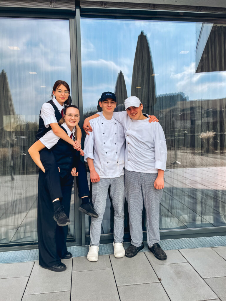 Man sieht vier Lehrlinge, zwei im Kochgewand und zwei im Restaurantgewand wobei eine die andere am Rücken trägt. alle wirken glücklich.