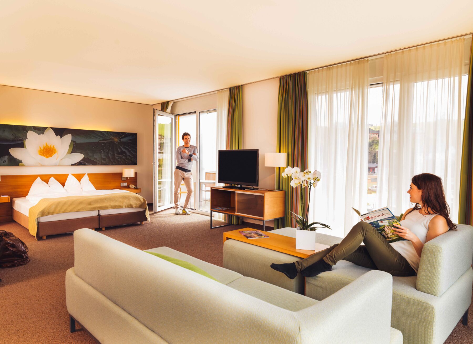 Hotelzimmer mit zwei Gästen. Die Dame sitzt auf der Couch und lest ein Magazin und der Mann lehnt an der Terrassentür mit Blick zur Frau. Im Raum befinden sich noch Bett und Fernseher.