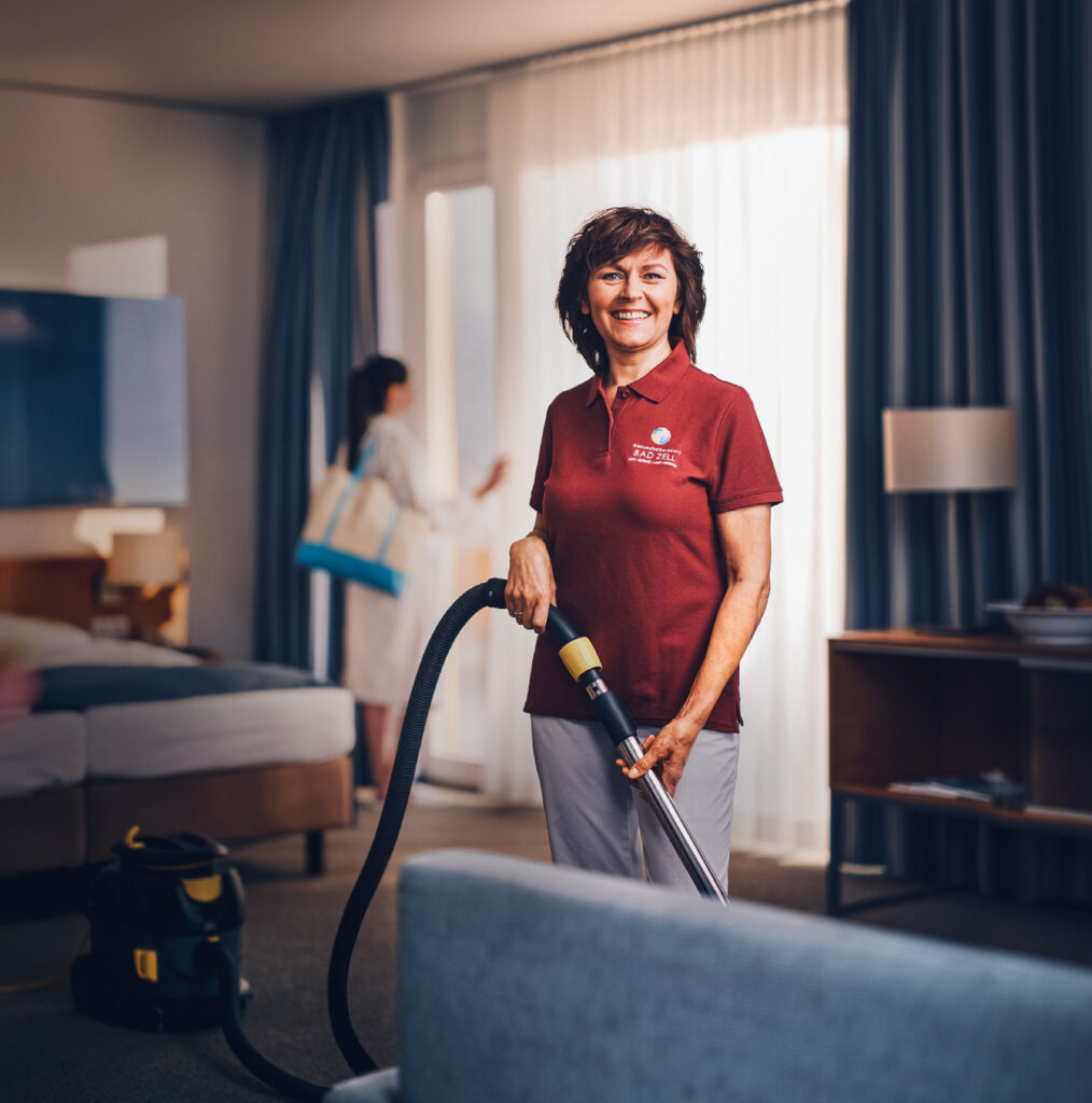 MAn sieht eine Reinigungskraft, welche einen Staubsauger in den HÄnden hält und sich in einem Gästezimmer befindet. Im Hintergrund ist ein Gast zu sehen, welcher den Ausblick genießt.