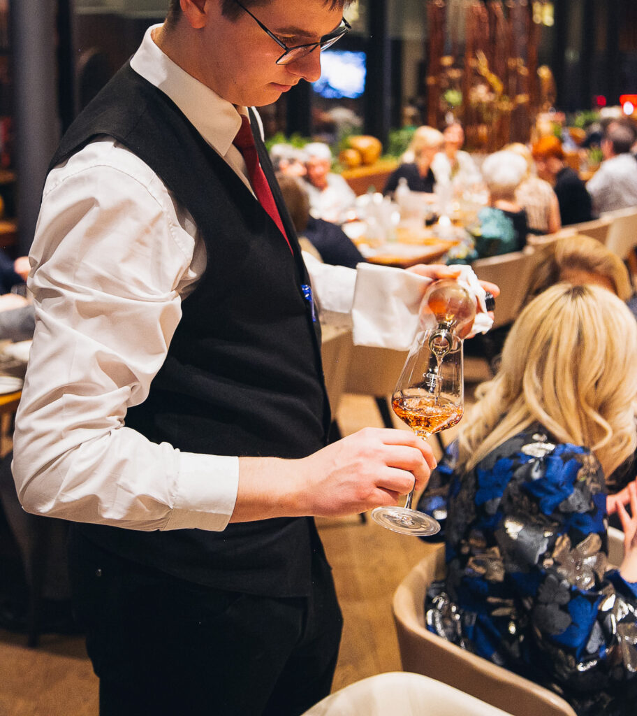 Man sieht eine Restaurantfachkraft beim Einschenken eines Glas Weines. Im Hintergrund Gäste, welche im Restaurant sitzen.