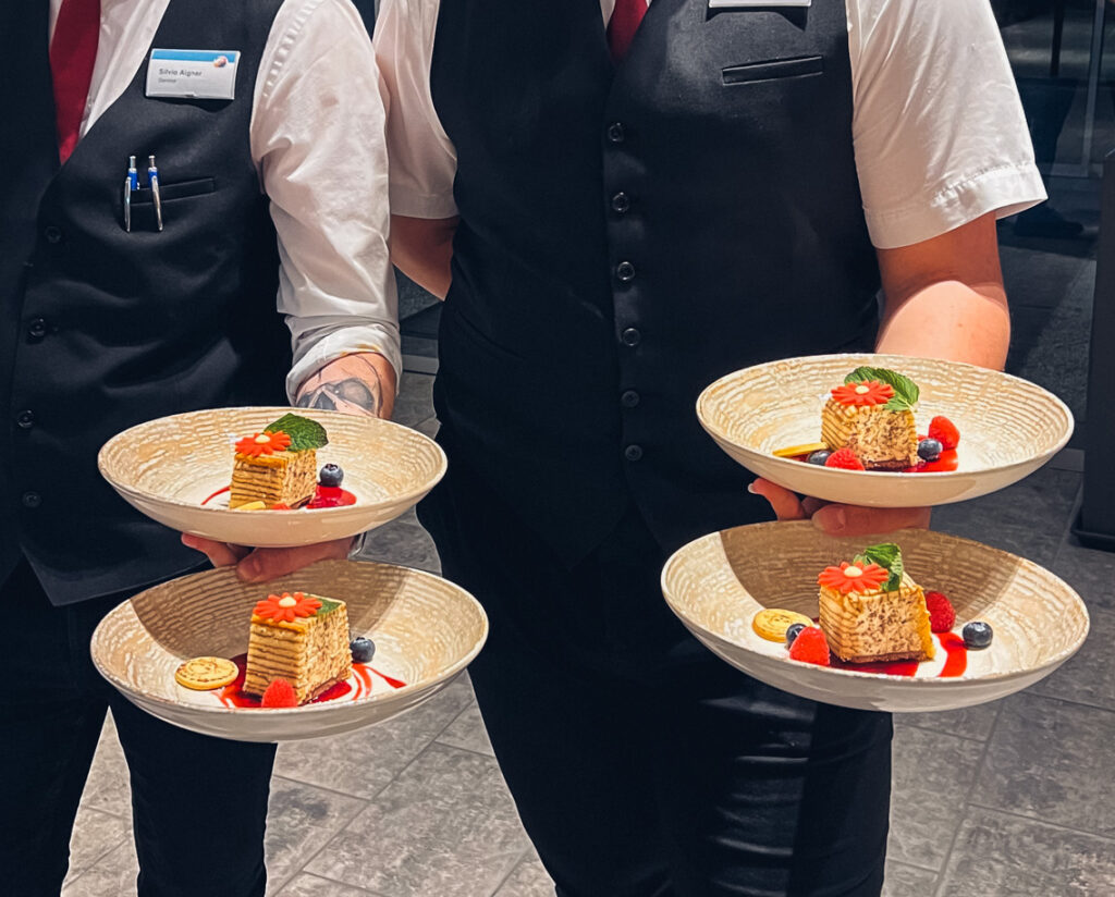 Man sieht zwei Chef de Rang mit jeweils zwei Tellern in der Hand mit einer süßen Nachspeise.