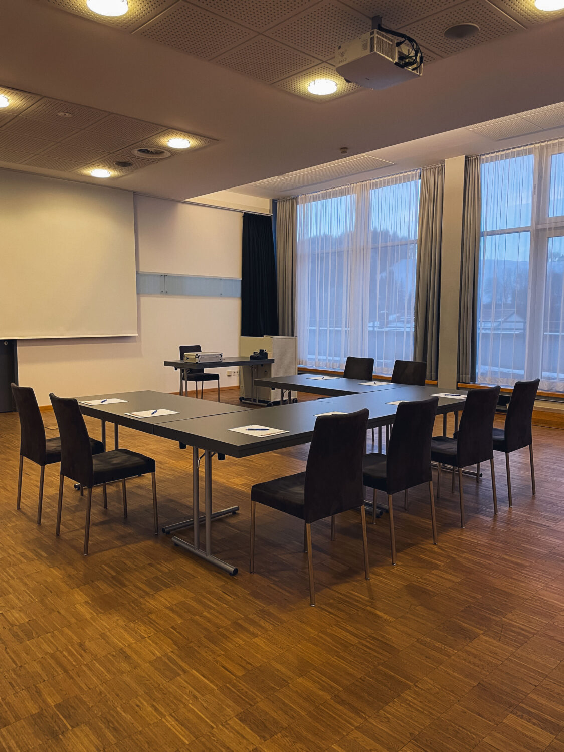 Seminarraum mit Tischen und Sessel in U-Form. Tische sind mit Blöcken und Kugelschreiber belegt. Davor eine Leinwand und Trainertisch mit Moderationskoffer.