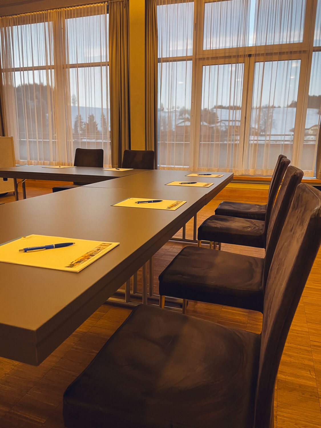 Aneinandergereihte Tische mit Stühlen im Seminarraum. Auf den Tischen befinden sich vor jedem Stuhl ein Block und Kugelschreiber.