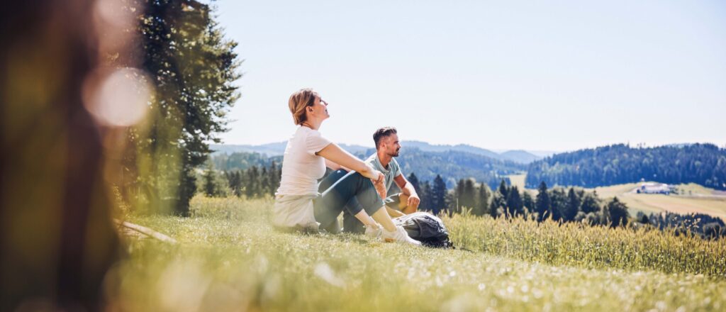 zwei Personen machen gerade eine Pause vom Wandern und sitzen in der Wiese und genießen die Sonne.