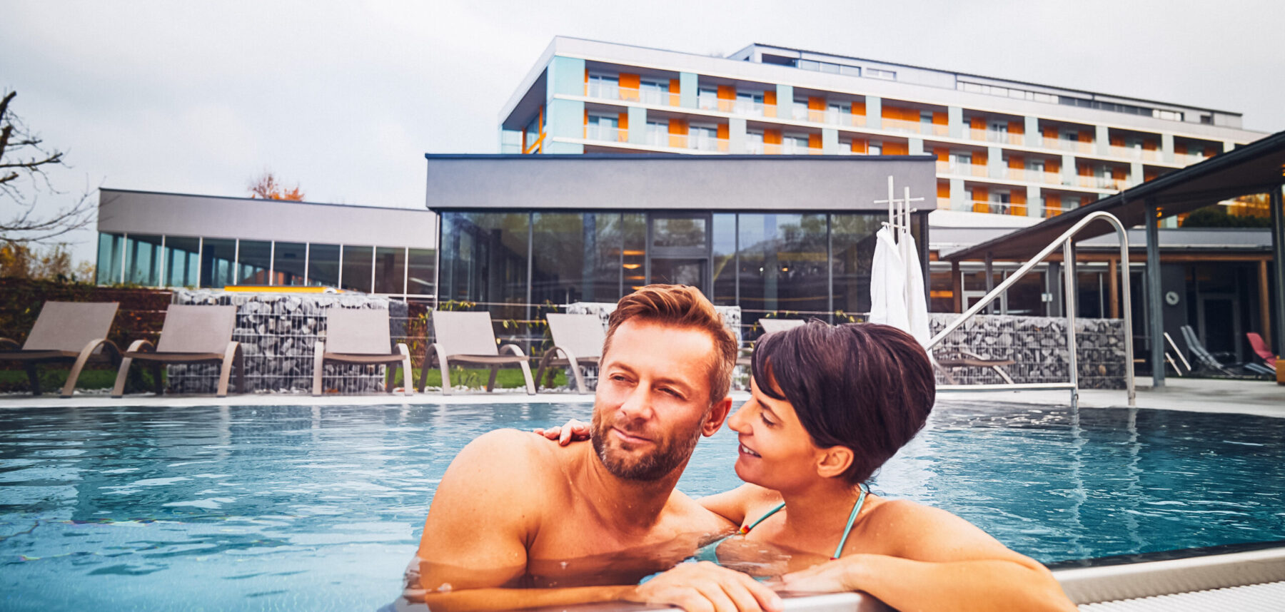 Ein Paar, dass sich am Seitenrand des Außenpools anlehnt und das Wetter genießt. Im Hintergrund das Hotel Lebensquell.