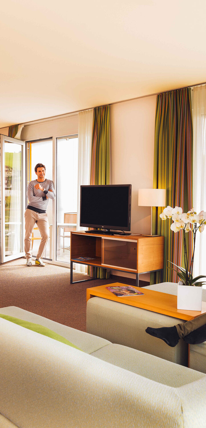 Hotelzimmer mit Fernseher und Couch. An der Terrassentür lehnt ein Mann.