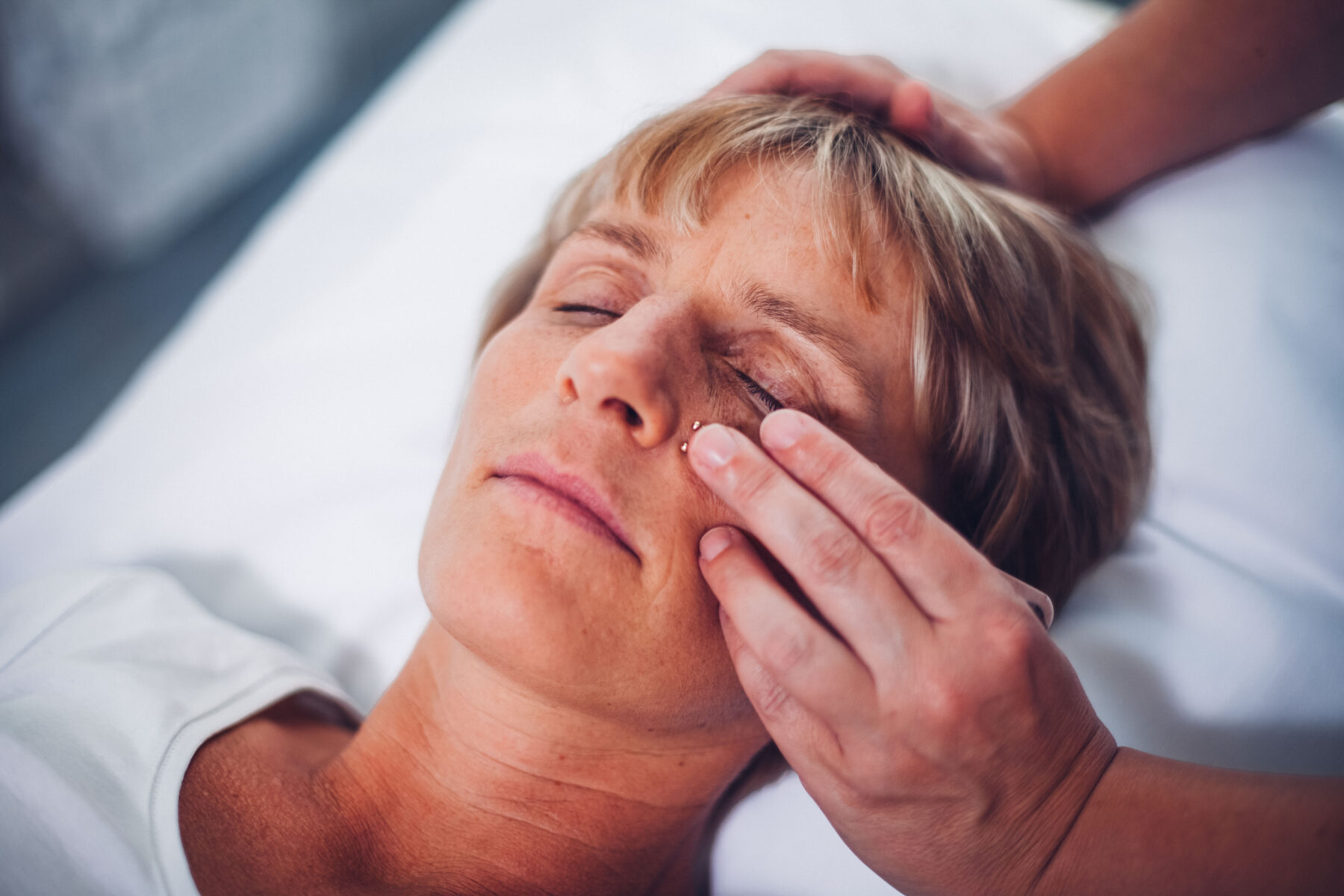 Frau liegt entspannt auf Behandlungsliege, Therapeutin platziert kleine Akupressurpflaster unter dem Auge.