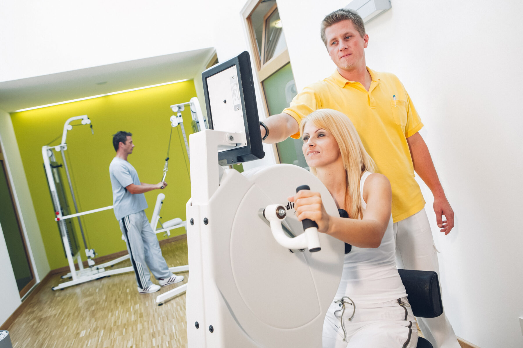 Physiotherapeut betreut Patientin an einem Trainingsgerät, während ein weiterer Patient im Hintergrund an einem anderen Gerät trainiert.