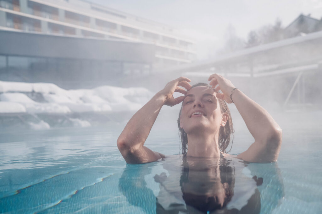 Eine Frau welche sich mit den Händen durch die Haare fährt und im Pool ist. Im Hintergrund das Hotel und Liegen, welche mit Schnee bedeckt sind und Dampf, der vom Becken aufsteigt.