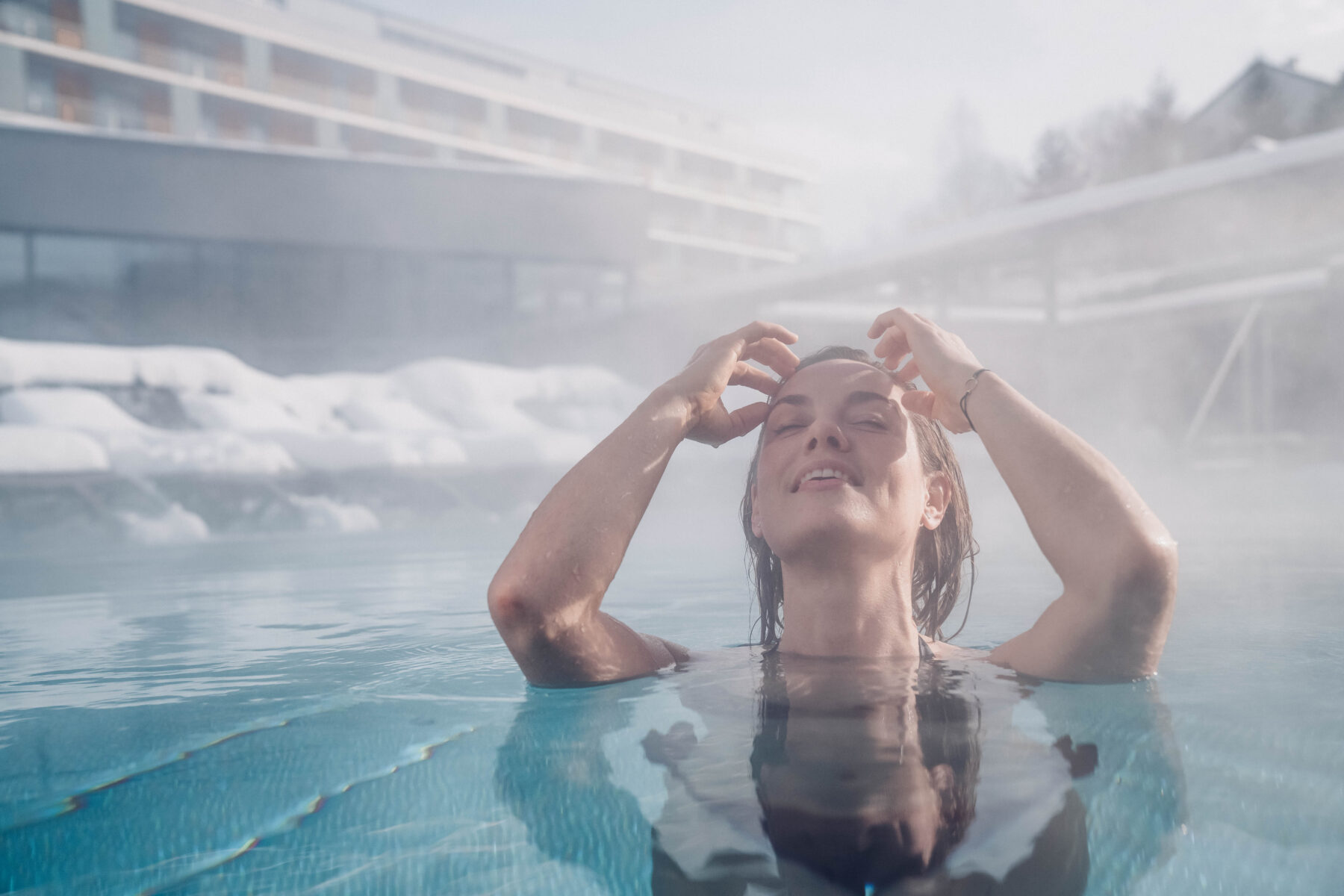 Eine Frau welche sich mit den Händen durch die Haare fährt und im Pool ist. Im Hintergrund das Hotel und Liegen, welche mit Schnee bedeckt sind und Dampf, der vom Becken aufsteigt.