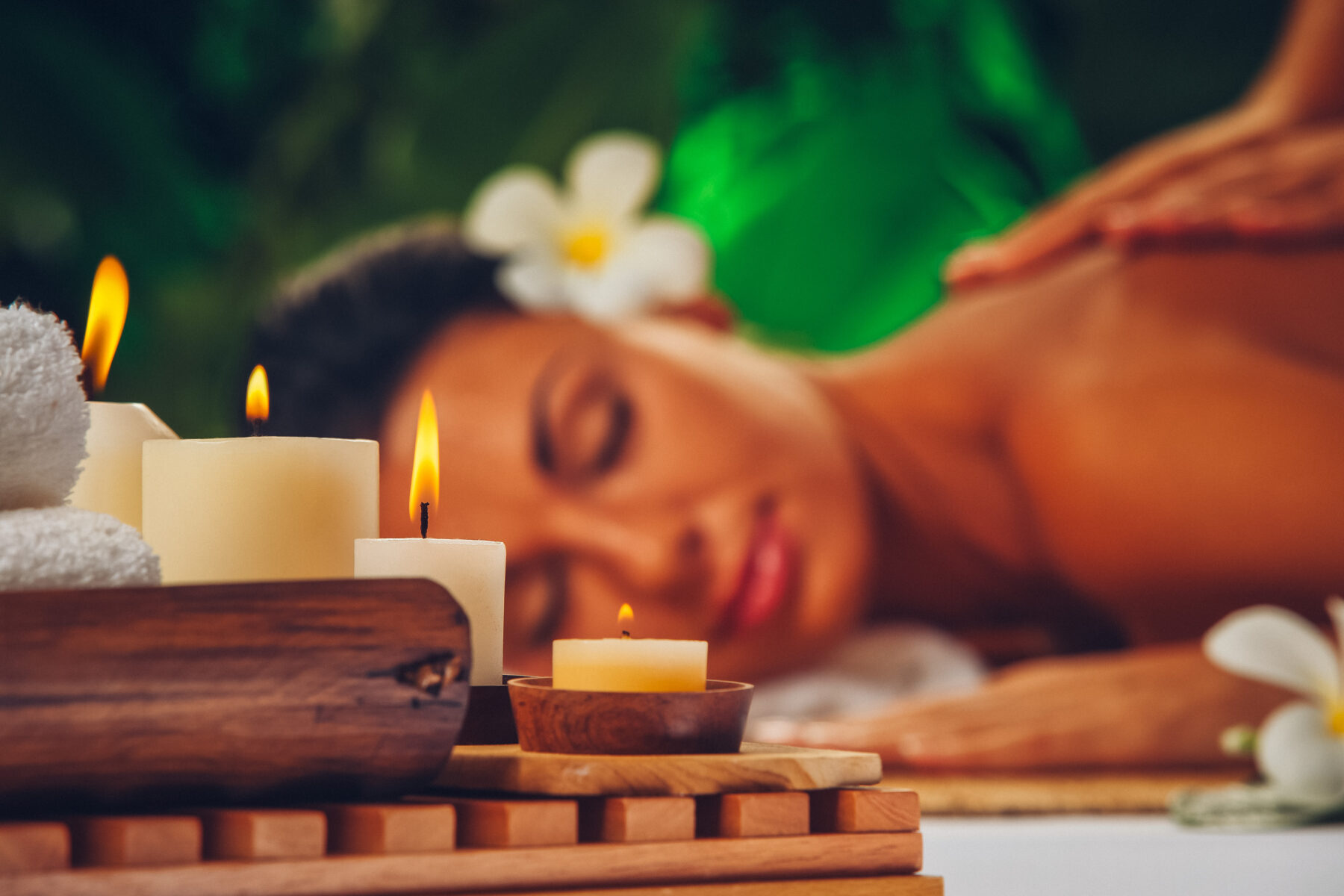 Eine Frau genießt eine entspannende Massage in einem tropisch inspirierten Spa-Ambiente. Im Vordergrund flackern sanft Kerzen, eingerahmt von weißen Handtüchern und Holzdetails.