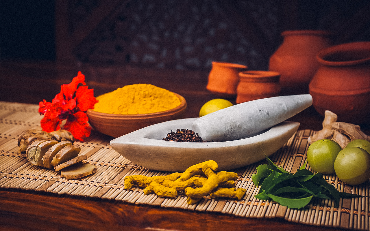 Verschiedene Kräuter und Gewürze, darunter Ingwer, Kurkuma, Nelken und grüne Früchte, liegen auf einer Unterlage mit einem Mörser und Stößel aus Marmor. Im Hintergrund sind Tontöpfe zu sehen.