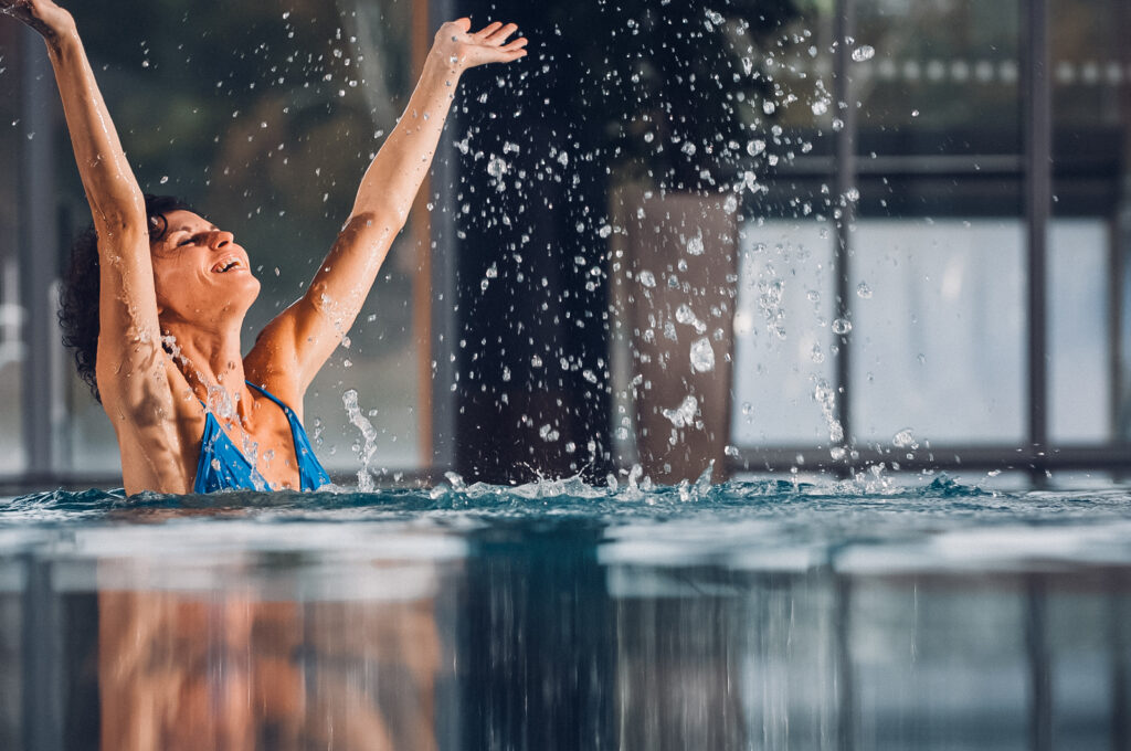 Frau in blauer Badekleidung hebt Arme freudig im warmen Innenpool.