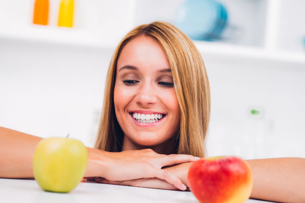 Eine lächelnde Frau schaut auf zwei Äpfel – einen grünen und einen roten – und wirkt dabei fröhlich und unbeschwert.