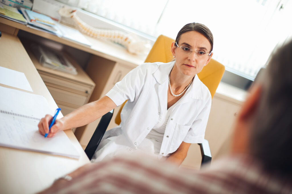 Arzt für Allgemeinmedizin in Ordination, welche gerade ein Gespräch führt und sich dabei Notizen macht.