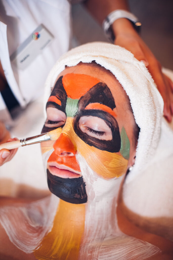 Auf dem Bild wird eine bunte Gesichtsmaske aufgetragen. Die Person entspannt mit geschlossen Augen und Handtuch am Kopf.