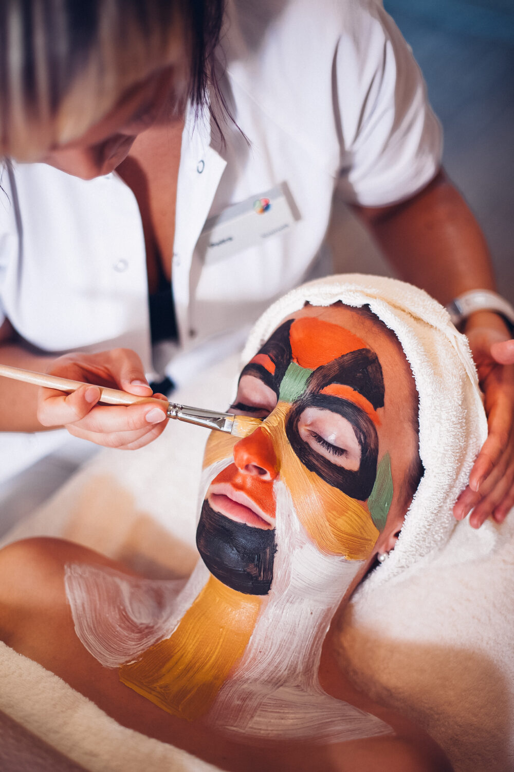 Auf dem Bild wird eine bunte Gesichtsmaske aufgetragen. Die Person entspannt mit geschlossen Augen und Handtuch am Kopf.