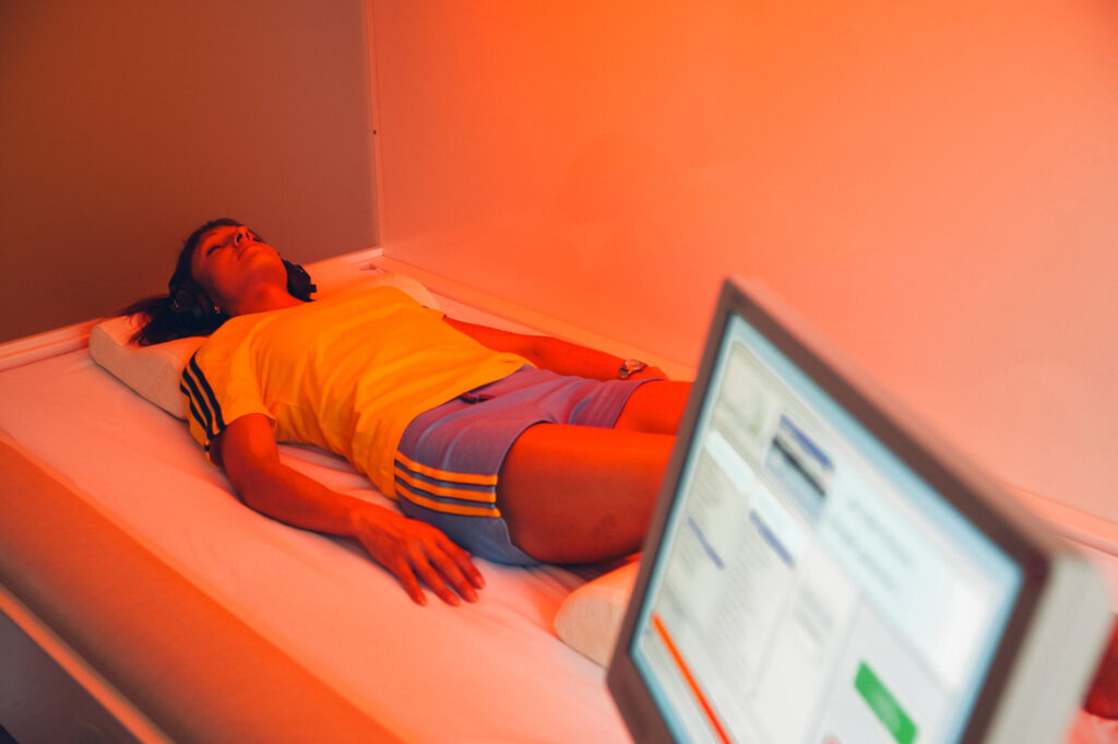 Eine Frau liegt entspannt in einem modernen Therapieraum unter warmem, rotem Licht, trägt Kopfhörer und genießt offenbar eine computergesteuerte Entspannungssitzung – eine Symbiose aus Technologie und Tiefenentspannung.