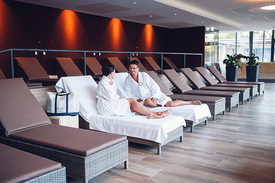 Ein Paar, das auf Liegen liegt und sich in die Augen sieht. Rundherum mehrere Liegen im Ruhebereich der Saunaoase.