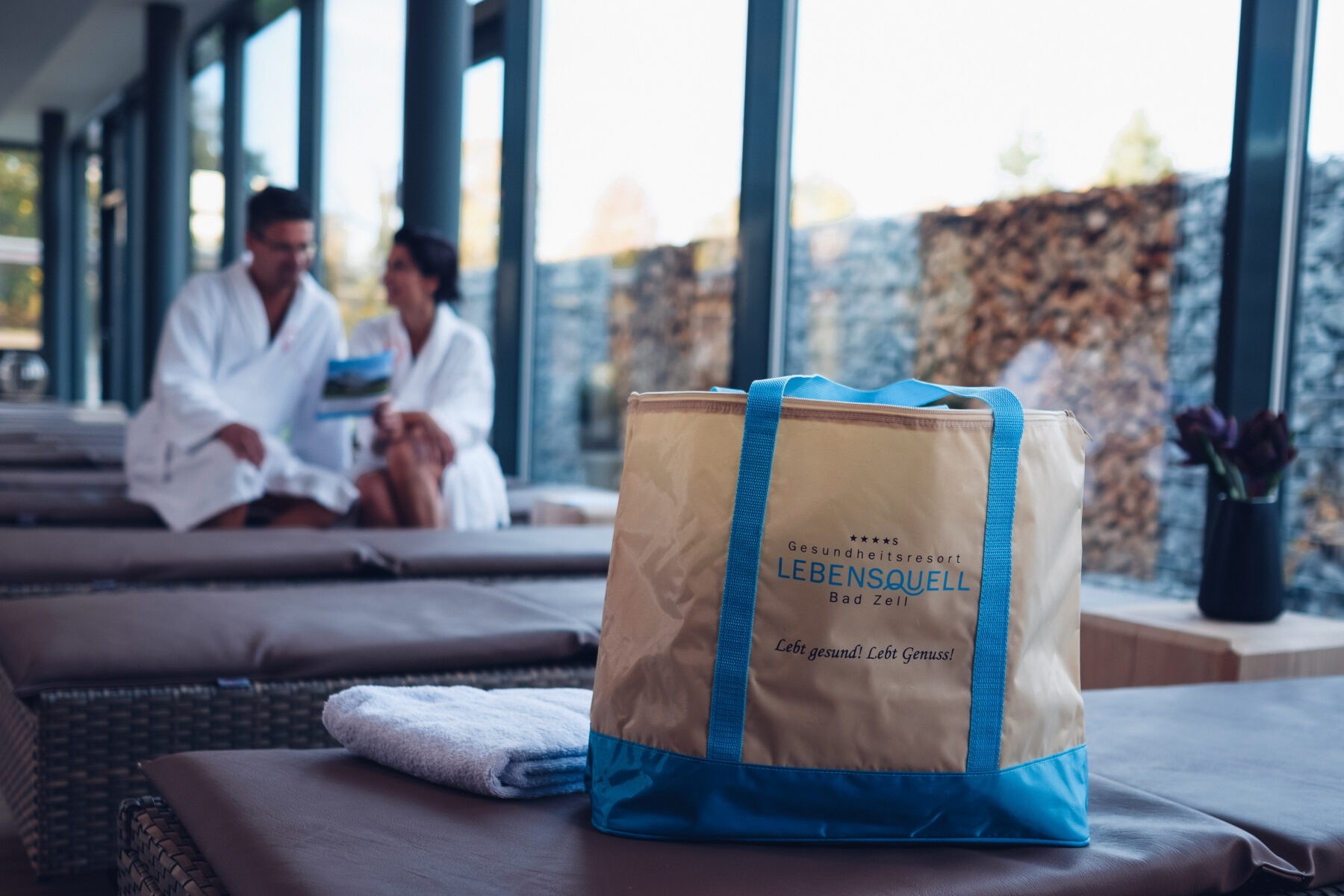 Im Vordergrund eine Lebensquell Badetasche, welche auf einer Liege steht. Im Hintergrund ein Paar sitzend auf einer Liege, am lesen von Broschüren.