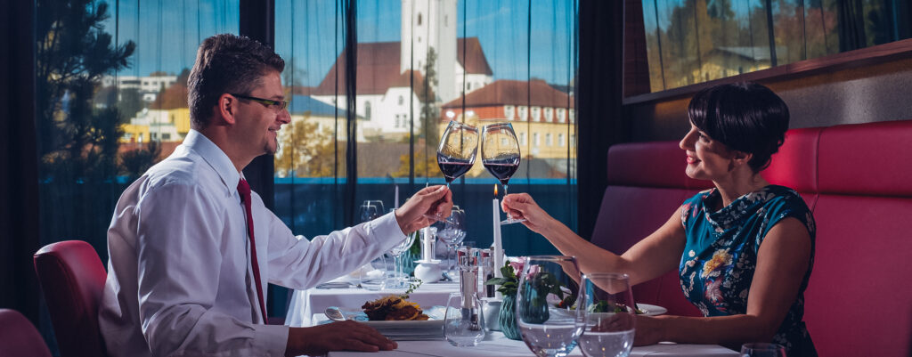Ein Paar, welches im Restaurant beim Abendessen ein Glas Wein genießen, mit welchen sie gerade anstoßen. Im Hintergrund das Restaurant mit einem Ausblick über Bad Zell.