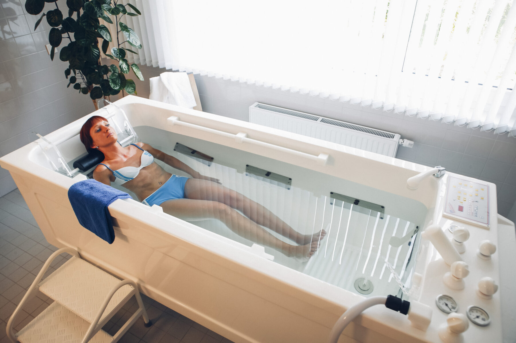 Eine Frau in Bikini liegt entspannt in einer modernen Hydrotherapie-Wanne in hellem Behandlungsraum.