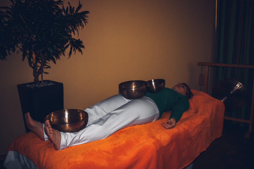 Frau liegt entspannt auf Liege, Klangschalen auf Körper für Klangschalenmassage platziert.