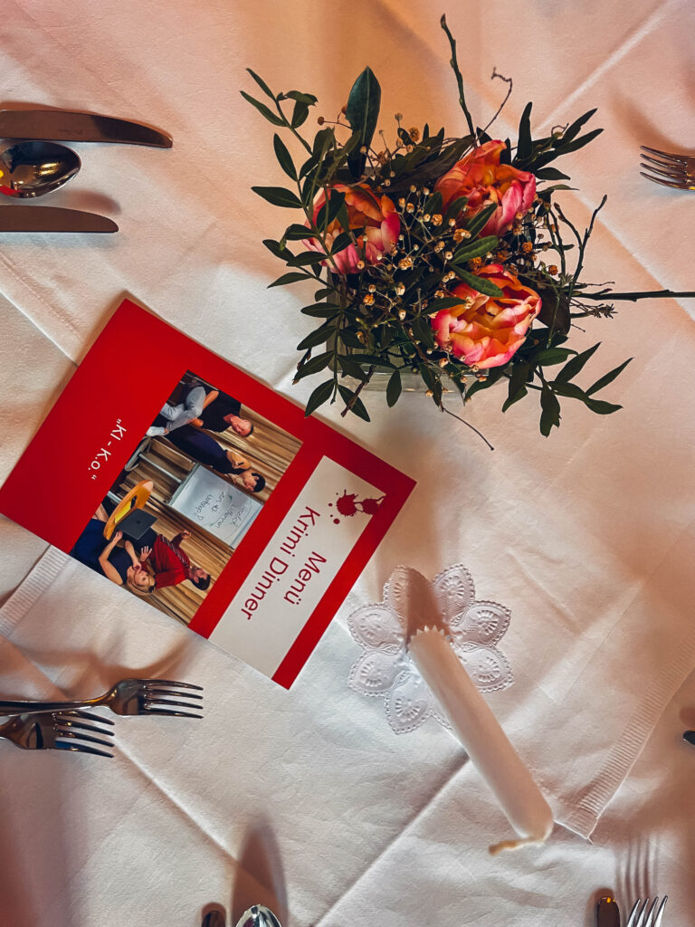 Man sieht einen gedeckten Tisch mit einer Kerze, einem Blumenschmuck und der Krimi-Dinner-Menü Karte.