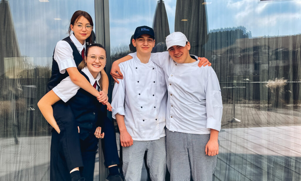 Man sieht vier Lehrlinge, zwei im Kochgewand und zwei im Restaurantgewand wobei eine die andere am Rücken trägt. alle wirken glücklich.