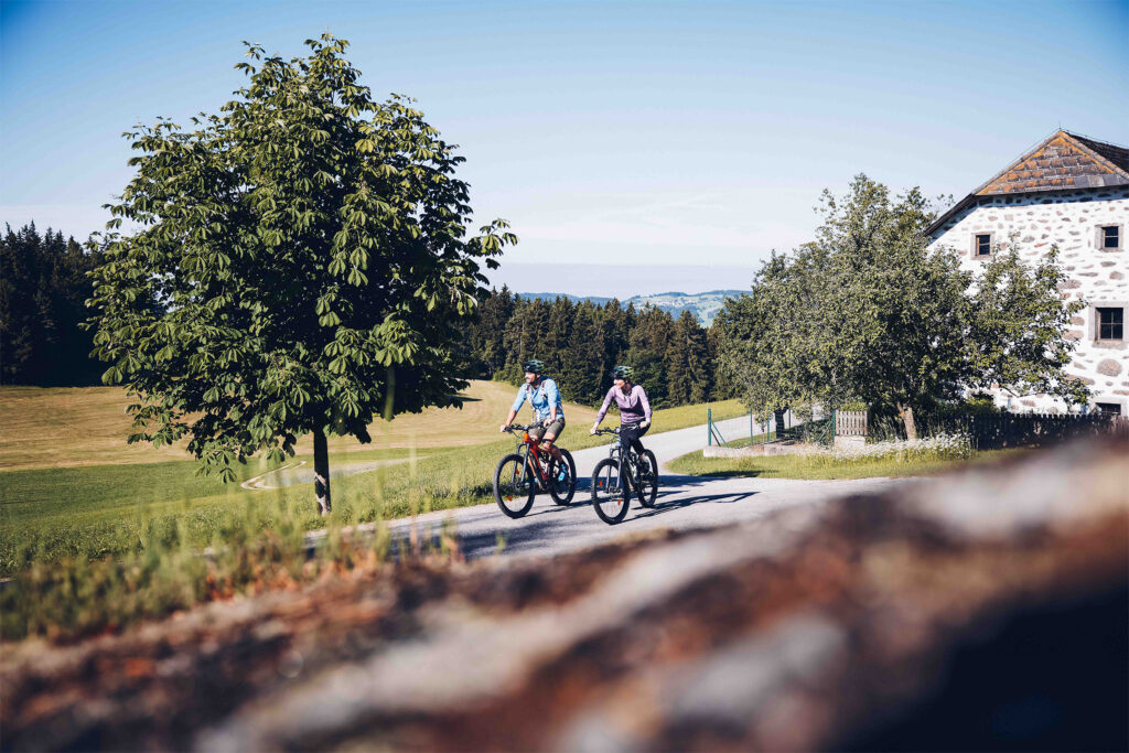 Ein Paar, welches gerade mit dem Rad bei einem Steinblashaus vorbeifährt, umgeben von Wald und Feldern. Im Hintergrund sind die Berge leicht zu erkennen.