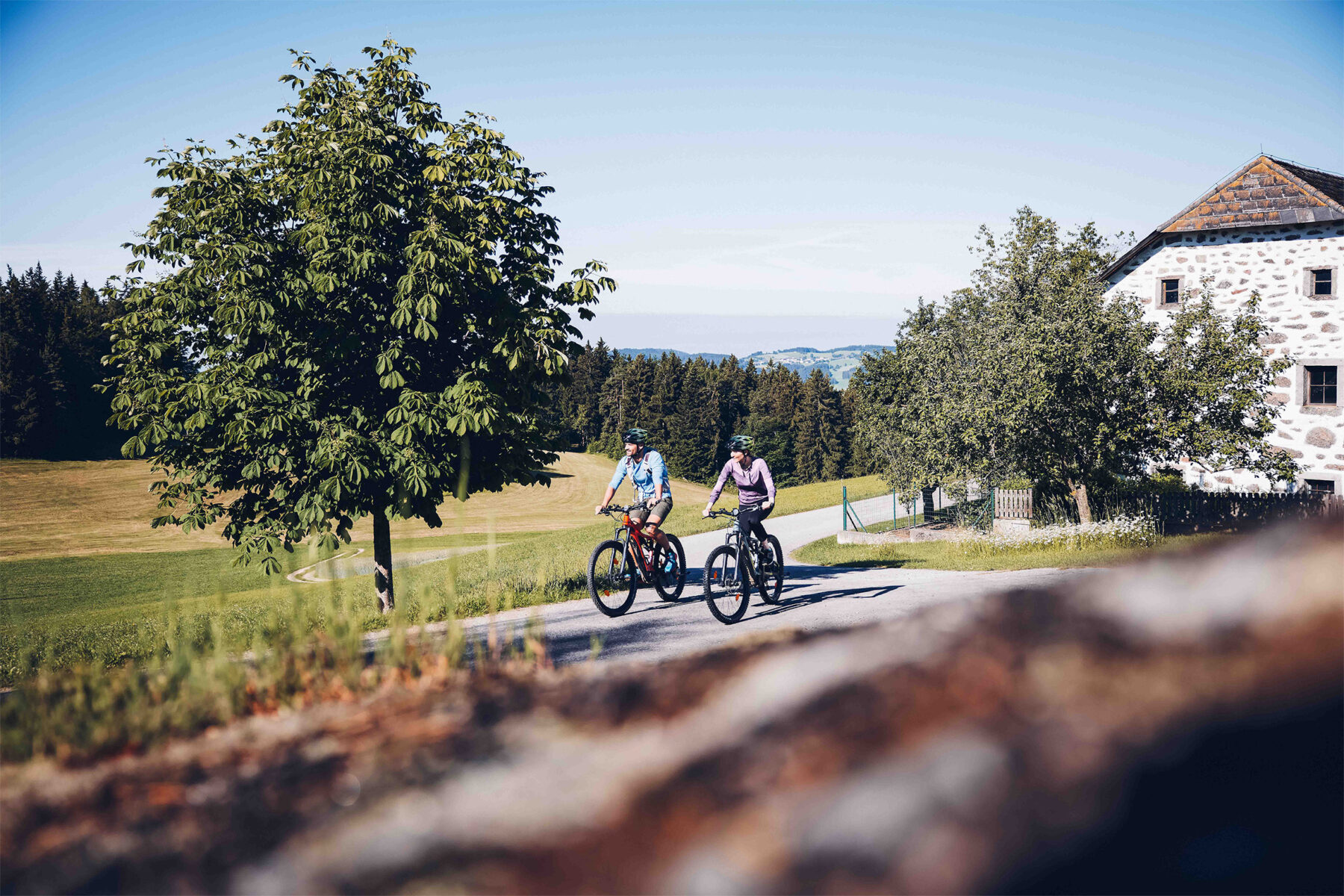 Ein Paar, welches gerade mit dem Rad bei einem Steinblashaus vorbeifährt, umgeben von Wald und Feldern. Im Hintergrund sind die Berge leicht zu erkennen.