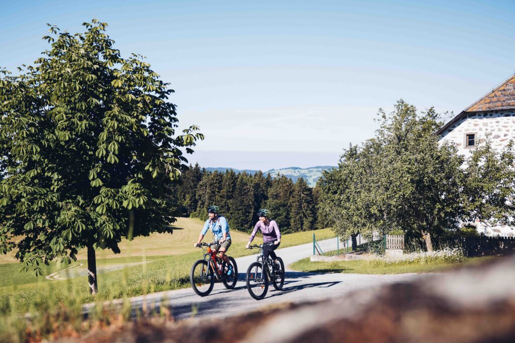 Ein Paar, welches gerade mit dem Rad bei einem Steinbloßhaus vorbeifährt, umgeben von Wald und Feldern. Im Hintergrund sind die Berge leicht zu erkennen.