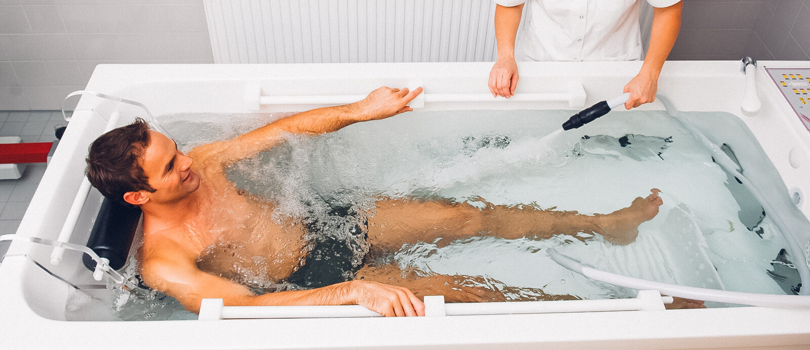Ein Mann in einer gefüllten Radonbadewanne. Ein Therapeut lässt mit einem Schlauch noch weiteres Wasser ein.