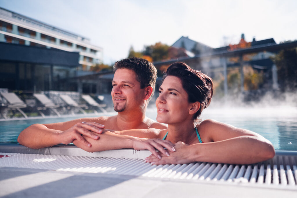 Ein Paar, dass sich am Seitenrand des Außenpools anlehnt und das Wetter genießt. Im Hintergrund das Hotel Lebensquell.