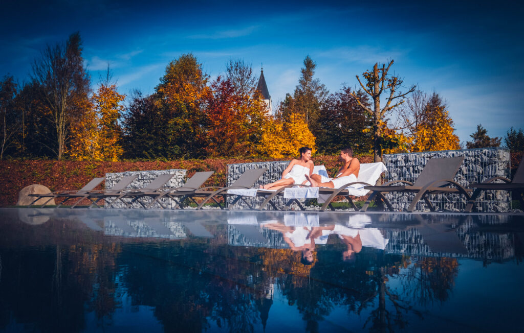 Ein Paar auf einer Liege vor dem Pool. Im Hintergrund die gelb/Rot gefärbten Bäume in herbstlicher Stimmung.