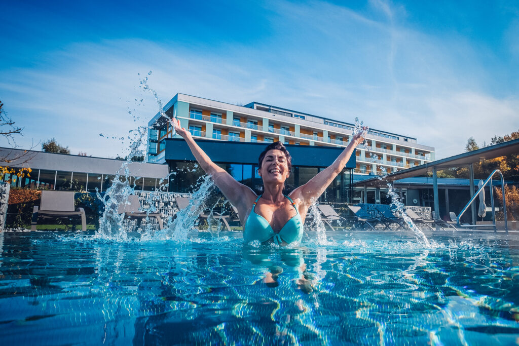 Glückliche Dame im Pool, welche vor Freude die Hände in die Luft hält und dabei mit dem Wasser spritzt. Im Hintergrund sieht man das ****s Hotel Lebensquell Bad Zell.