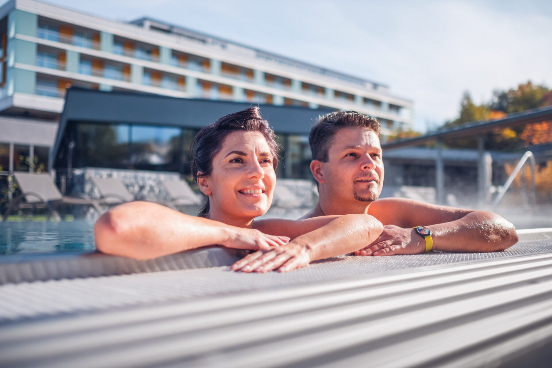 Ein Paar, dass sich am Seitenrand des Außenpools anlehnt und das Wetter genießt. Im Hintergrund das Hotel Lebensquell.