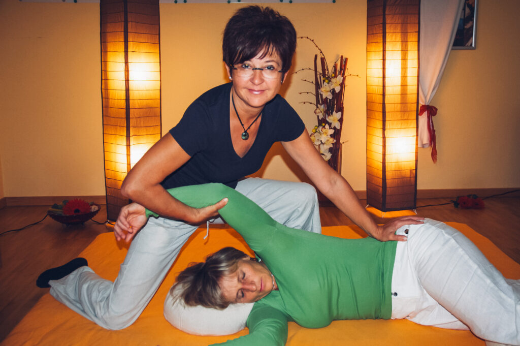Therapeutin führt Dehnübung mit liegender Frau in entspannter, warm beleuchteter Umgebung durch.